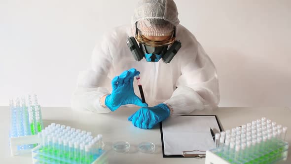 Doctor in a White Protective Overalls and a Respirator Looking on a Blood Sample in a Test Tube and