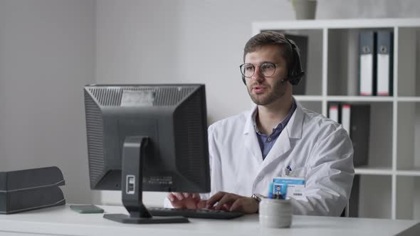 Male General Practitioner in White Coat Sitting at Desk in Doctor's Office and Scrolling Computer