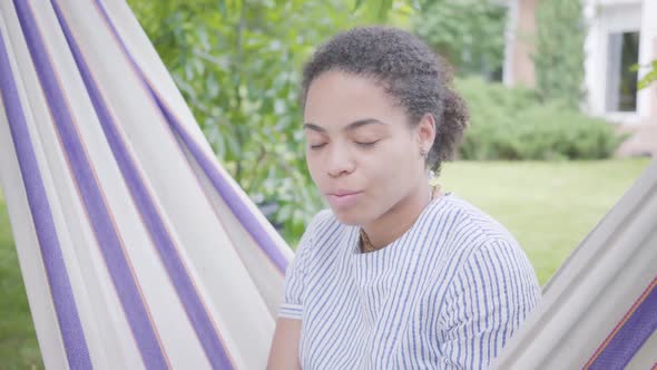 Portrait of Cute Young Smiling African American Woman Sitting in the Hammock, Relaxing in the Garden