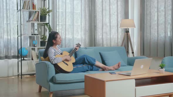 Asian Woman With A Laptop And Smartphone Relaxing Leaning On Sofa To Sing And Play A Guitar