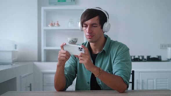 Young Man in Headphones Playing App Game on His Smartphone While Sitting on Kitchen Table at Home