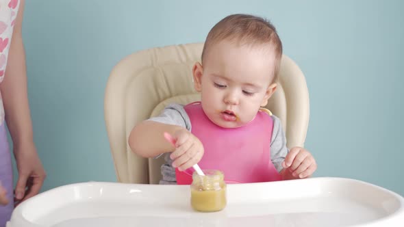 Mom teaches a newborn baby to eat food with a spoon on his own. Baby eats sitting on a feeding chair