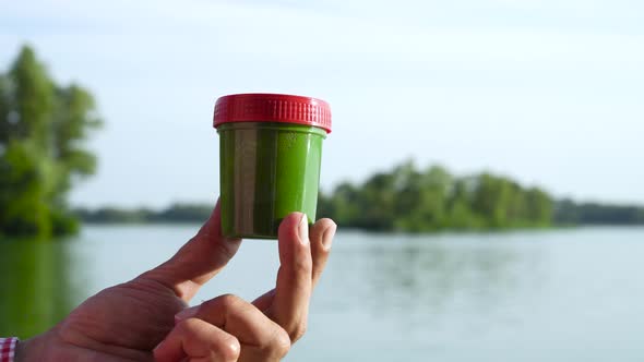 Closeup Male Hand Shaking Container with Green Algae with River on Background