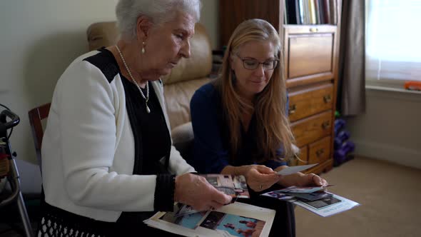 Elderly gray haired woman and blonde, mature woman look at photos in a photo album.