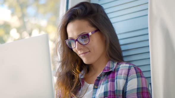 Young Woman Dressed Casually Working on Laptop While Sitting on Window Sill at Home