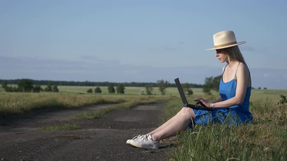 Girl in blue dress with laptop computer sit on country road in summer.