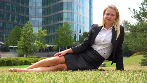 A Young Beautiful Businesswoman Sits on Grass in a Park and Smiles at the Camera - Office Buildings