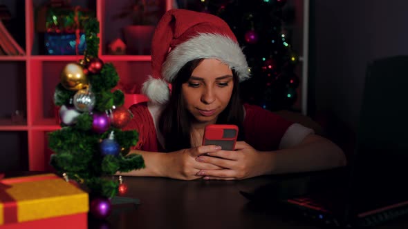 Young Woman in Santa Costume Using Smartphone in Office Sitting in Armchair in New Year's Atmosphere