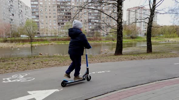 Little Boy Riding a Scooter on a City Bike Path in Early Spring