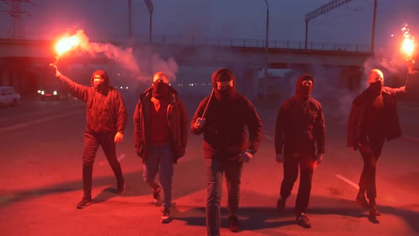 Group of Young Men in Balaclavas with Red Burning Signal Flare Walking on the Road Under the Bridge