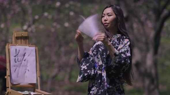 Gorgeous Asian Young Woman in Kimono Dancing with Fans in Spring Sakura Garden Smiling As Blurred