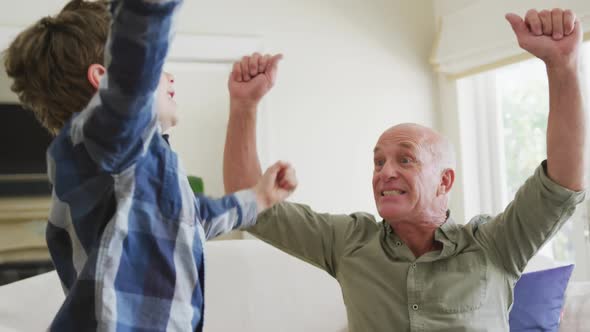 Happy caucasian grandfather and grandson jumping and raising hands in living room