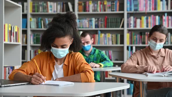 Multiracial Students in Protective Medical Masks Sit at Tables at a Safe Distance in the Library