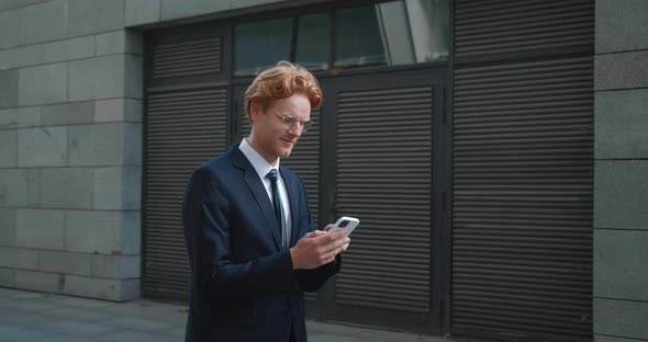 Young Caucasian Male Businessman Typing on Smartphone While Walking Along Modern Business Building