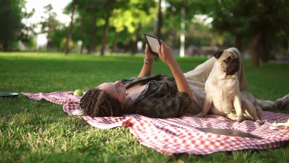 Woman Laying on Plaid on Lawn in a Park and Reading Ebook While Little Pug Sitting Next to Her