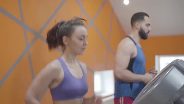 Side View of Slender Young Caucasian Woman Running on Treadmill and Talking with Blurred Middle