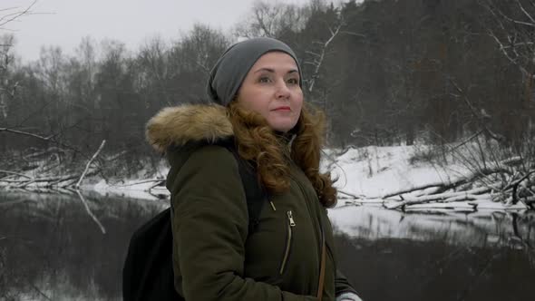 a Girl with Red Hair in a Dark Green Jacket and a Backpack Stands Near the River and Looks Around