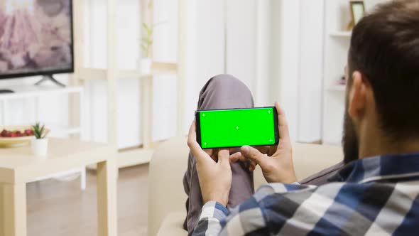 Caucasian Man Holding a Green Screen Smartphone in His Hands While Looking at the TV