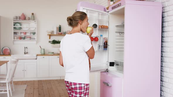 Young Girl in Home Clothes is Eating Apple Opening Fridge Taking Out Salad Leaves Leaving for