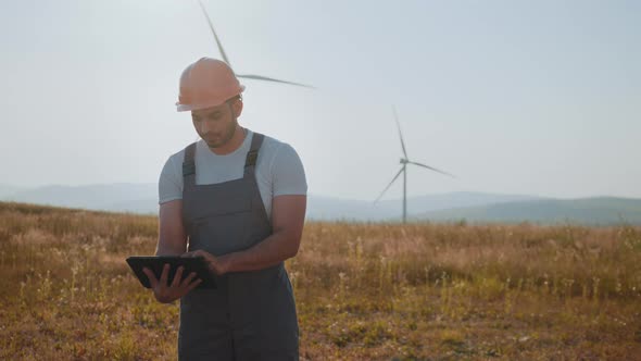 Portrait of Focused Indian Man Standing on Field with Wind Turbines and Using