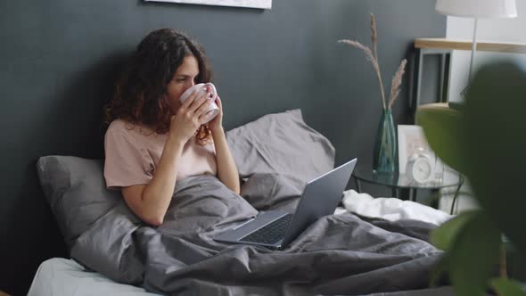 Woman Drinking Coffee and Typing on Laptop in Bed