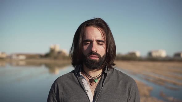 Long haired man standing near lake with closed eyes