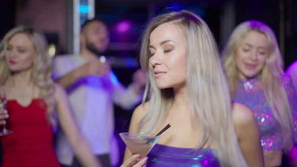Portrait of Young Slim Charming Woman with Dyed Blond Hair and Green Eyes Dancing with Cocktail