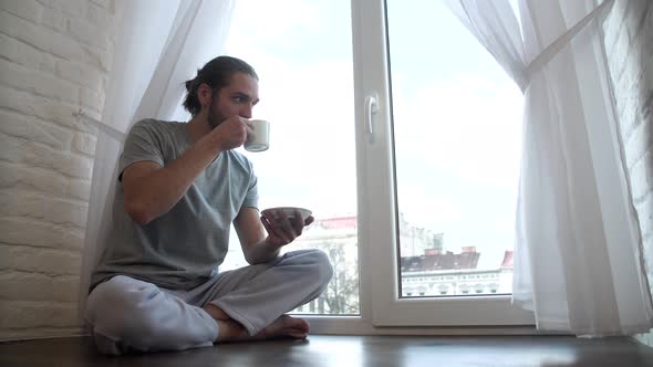 Man In Pajamas Drinking Coffee And Sitting On Window