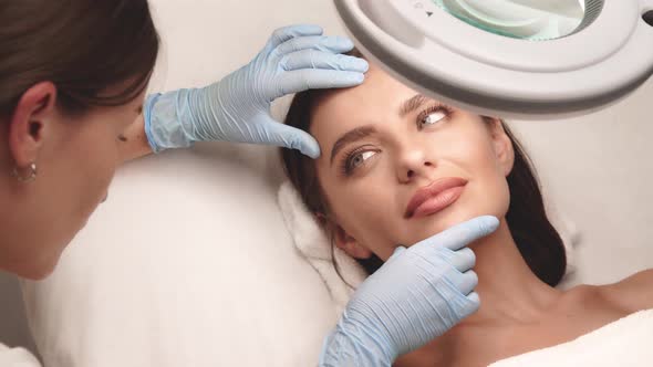 Cosmetologist Checking Female Cheeks Under Lamp at Beauty Clinic