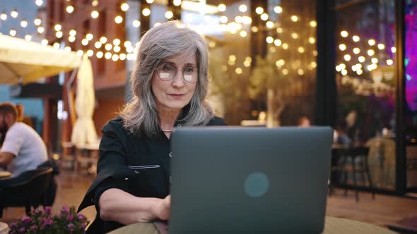 Elegant Mature Grayhaired Business Woman Sitting and Working in the Evening in a Cafe Against the