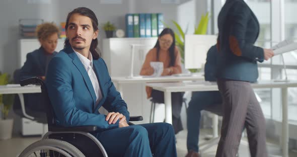 Sad Disabled Businessman Sitting in Wheelchair in Office with Colleagues Working on Background