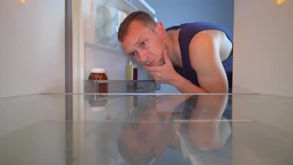 man looks into an empty refrigerator and takes out bottle of alcohol. Alcoholism