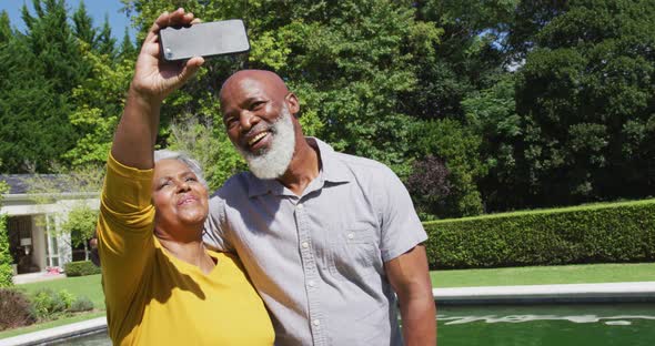 Happy senior african american couple using smartphone taking selfie by swimming pool in sunny garden