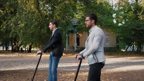 Two Handsome Young Businessman Riding an Electric Scooters in City Park During a Break Slow Motion