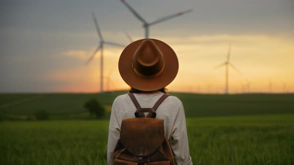 Portrait of a young girl in a hat and with a backpack looking at a wind farm at sunset