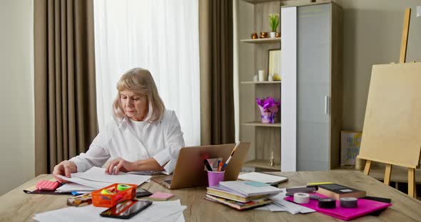 Senior Businesswoman Working on Paperwork in a Home Office Arranging Papers on Her Desk with Laptop