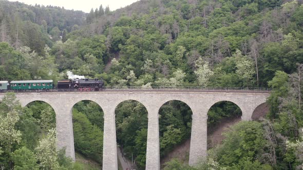 Train with a steam locomotive driving over a stone viaduct in a valley.