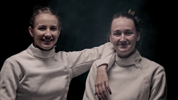 Two Young Smiling Women Fencers Standing in the Studio and Looking in the Camera