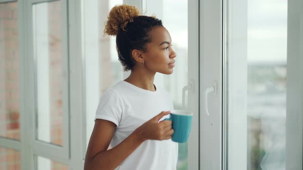 Happy Mixed Race Woman Is Looking Out of the Window and Drinking Coffee Standing Near Large Window