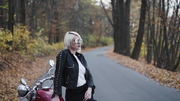 Portrait of Blonde in Sunglasses and Leather Clothes Poses at Motorbike in Park