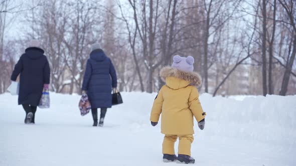 toddler 12-17 months old in yellow winter clothes looks after two middle-aged women