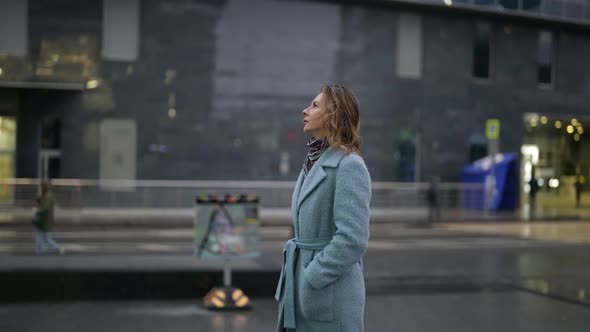 a Woman in a Light Coat Has Hands in Pockets and is Walking Against the Background of a Blurry