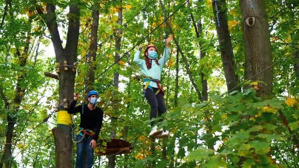 Little girl and instructor in medical masks and safety equipment engaged climbing rope park. 