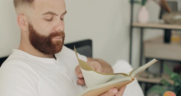 Close Up View of Concentrated Young Man Reading Novel While Having Breakfast in Bed. Handsome