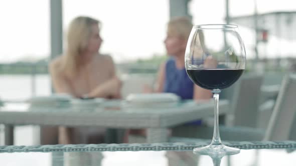Glass with Red Wine Standing at the Table in Outdoor Cafe and Two Blurred Blond Caucasian Women