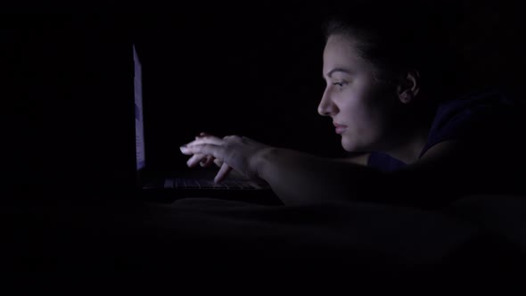 Attractive Young Happy Woman Uses a Mobile Computer While Lying on a Bed at Night. Isolated Light 