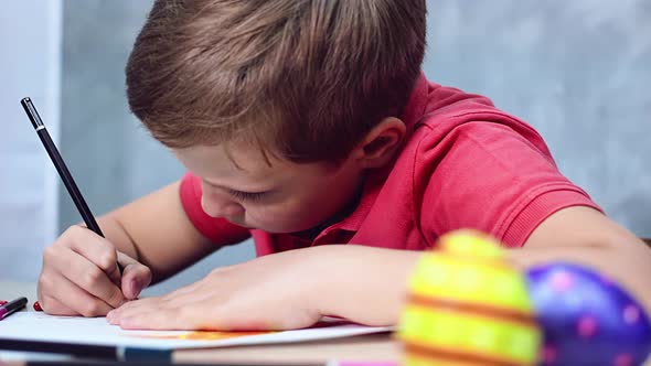 A boy is drawing an Easter drawing with coloured pencils on a white sheet of paper