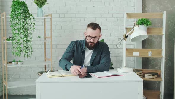 Bearded Guy Learn Online Courses Using a Tablet and Write Notice in His Notebook