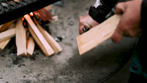 man cutting wood shavings of a log besides campfire