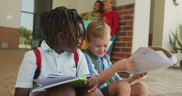 Video of happy diverse boys holding books and talking in front of school